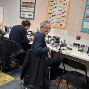 Smiling attendee at a soldering station during a Guitar Amp Academy pedal building workshop, with others building pedals behind him