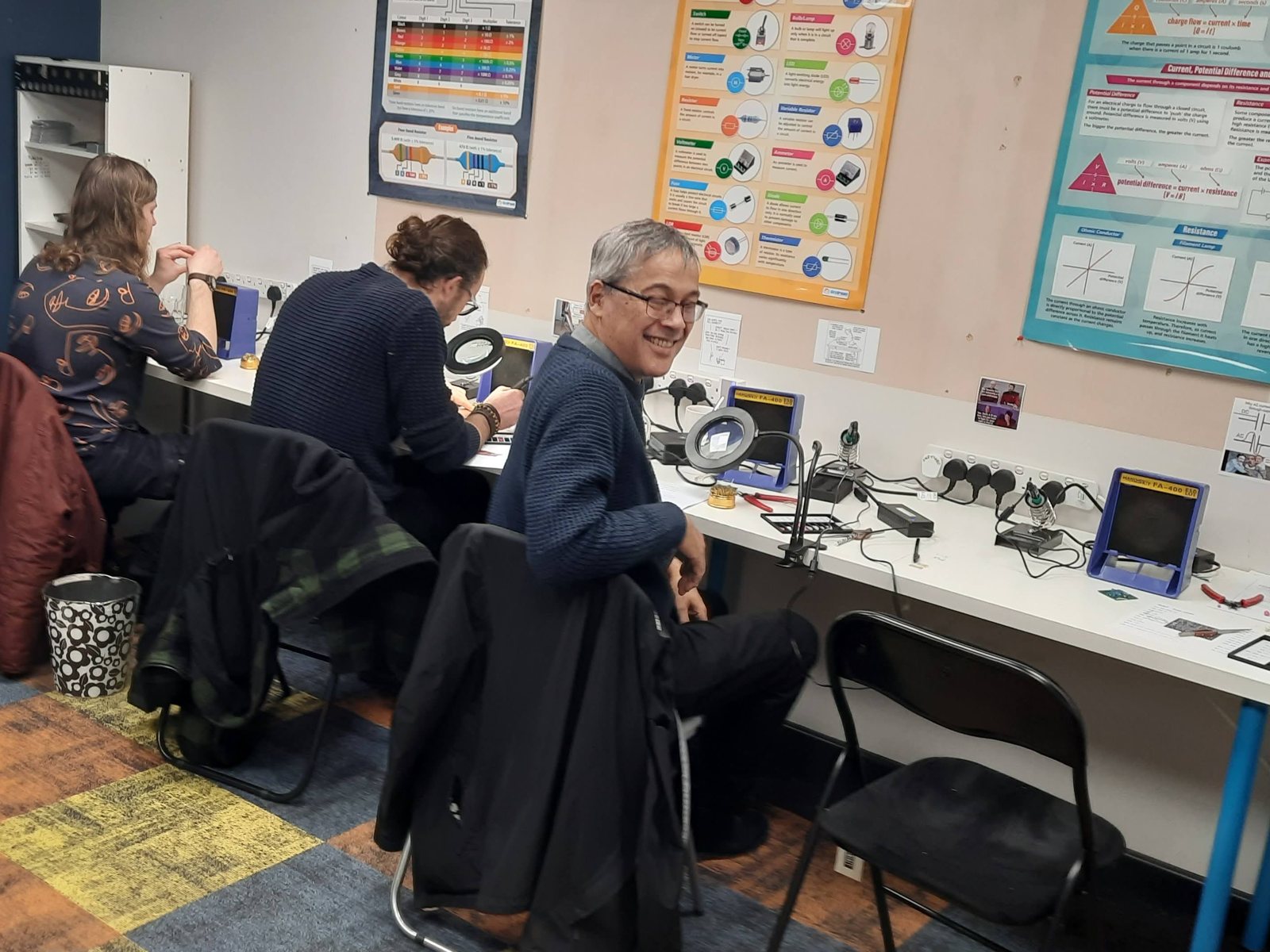 Smiling attendee at a soldering station during a Guitar Amp Academy pedal building workshop, with others building pedals behind him