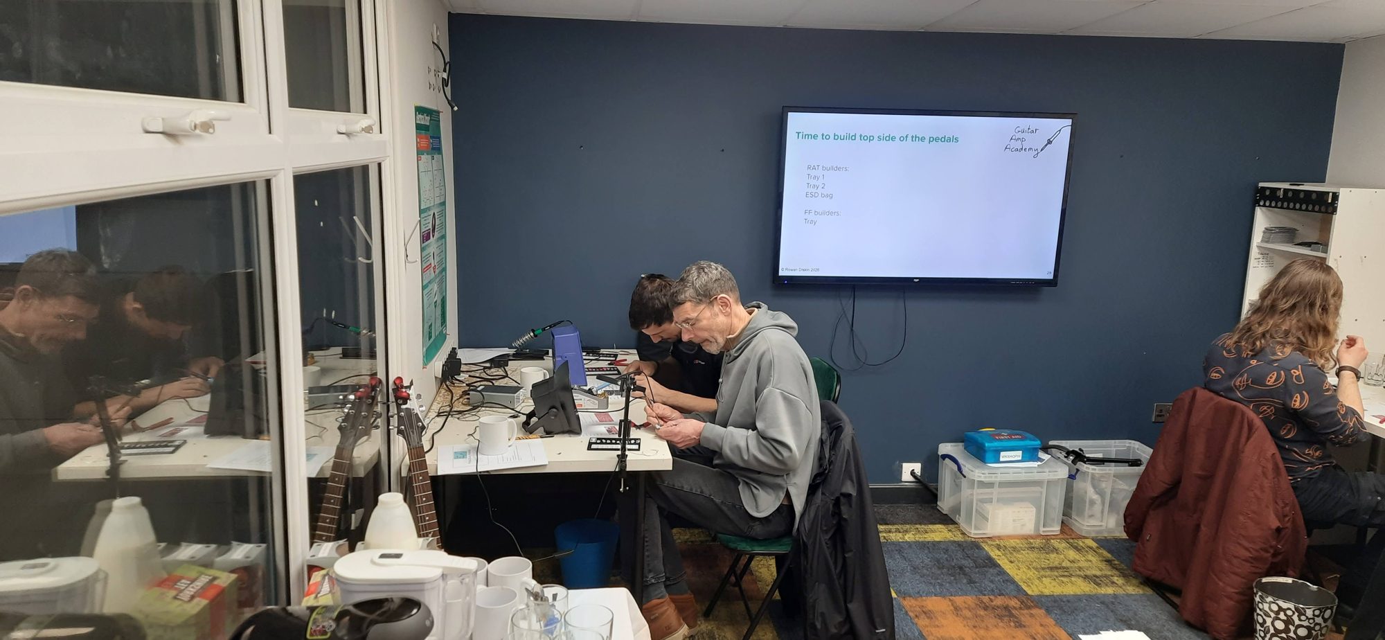 Attendees soldering pedal circuits while a screen shows instructions for RAT and Fuzz Face builders