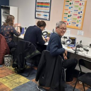Three attendees at soldering stations building guitar effects pedals, with electronics posters on the wall