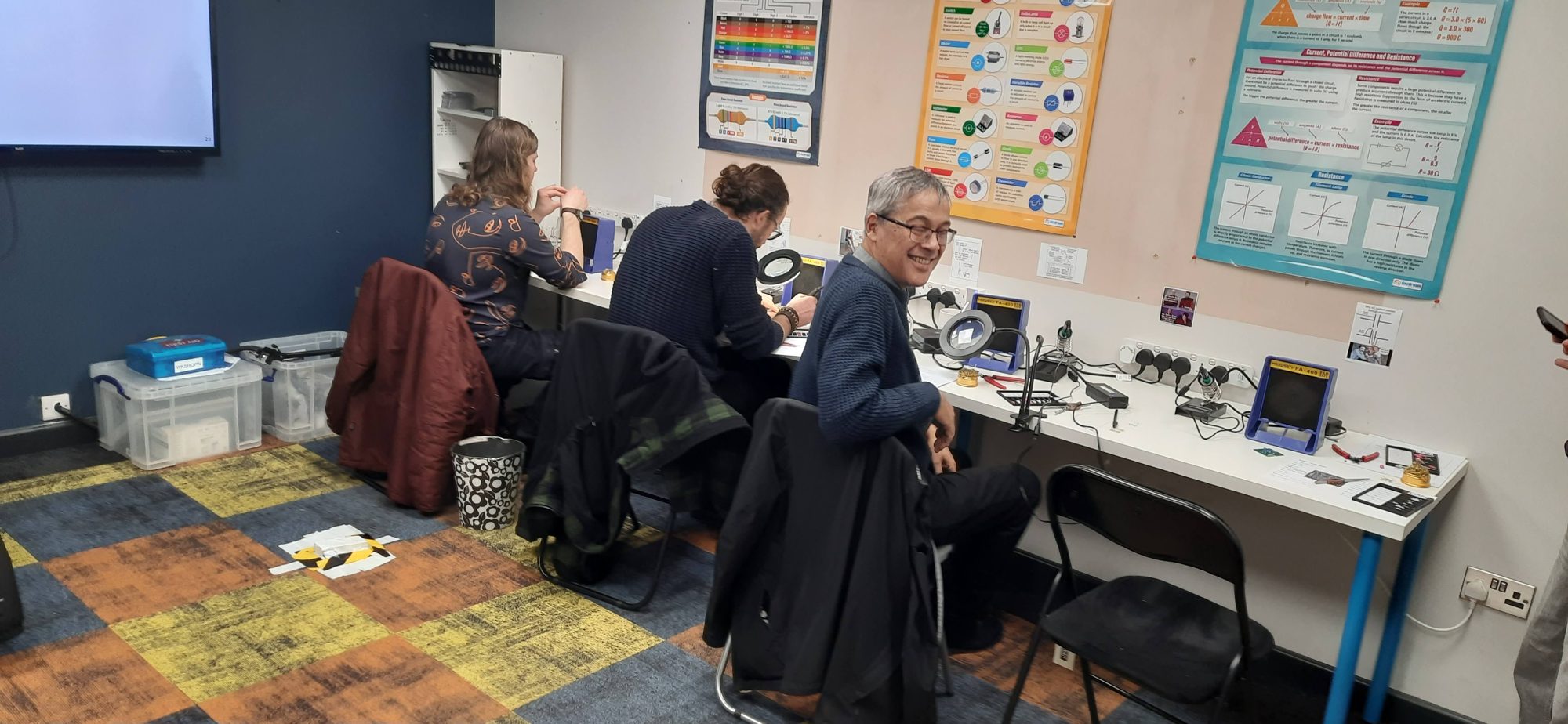 Three attendees at soldering stations building guitar effects pedals, with electronics posters on the wall