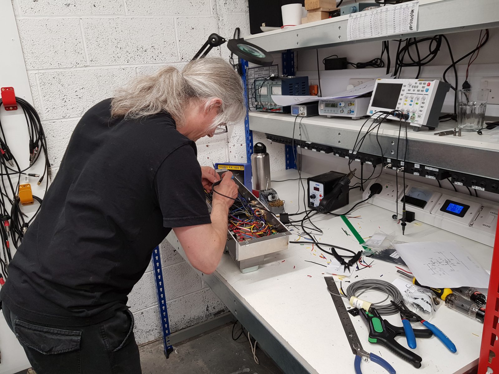 Builder soldering point-to-point wiring inside an aluminium valve amp chassis, with test equipment on the shelves behind
